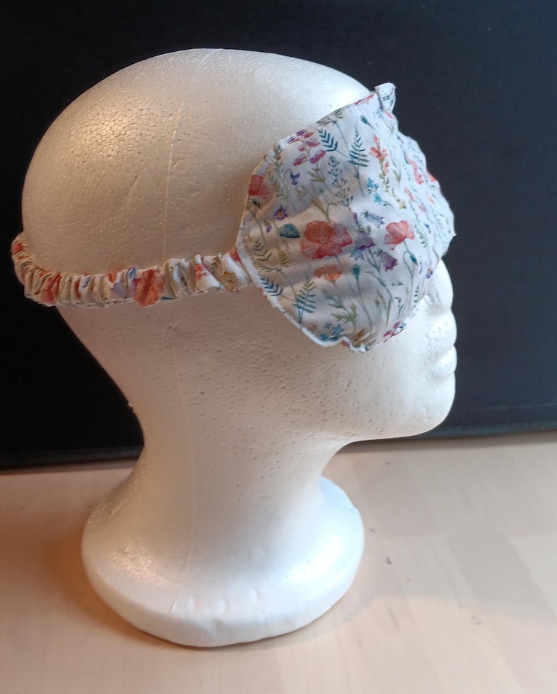 May include: A floral sleep mask with a white base, adorned with red, orange, and blue flower patterns. The mask includes an elasticated band. This sleep mask is designed to block out light for improved sleep.