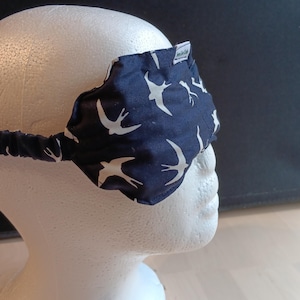 May include: A navy blue sleep mask featuring a white bird design. The mask is made of fabric and has an elasticated band. The mask is on a white mannequin head. The mask has a small tag with text.