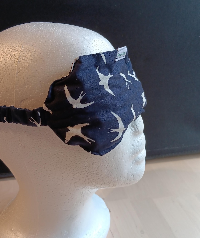 May include: A navy blue sleep mask featuring a white bird design. The mask is made of fabric and has an elasticated band. The mask is on a white mannequin head. The mask has a small tag with text.