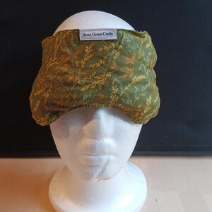 May include: A green and gold patterned eye mask with the text "Sewn Green Crafts" on a white label. The mask is placed on a white mannequin head. The mask has a leaf and branch design.