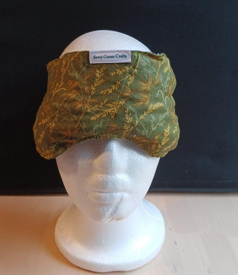 May include: A green and gold patterned eye mask with the text "Sewn Green Crafts" on a white label. The mask is placed on a white mannequin head. The mask has a leaf and branch design.
