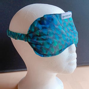 May include: A blue and green sleep mask with a scale pattern, worn on a white mannequin head. The mask has an elastic band and a small white tag that reads "Sewn Green Crafts".
