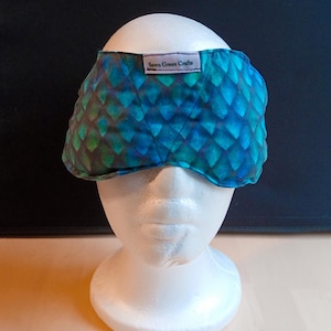 May include: A quilted eye mask with a blue and green scale pattern, displayed on a white mannequin head. The label on the mask reads "Sewn Green Crafts".