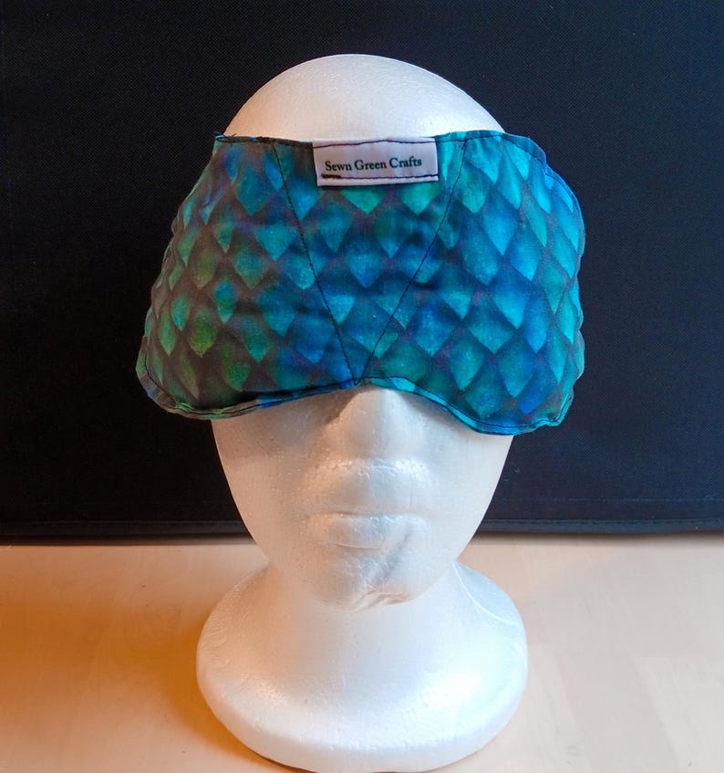 May include: A quilted eye mask with a blue and green scale pattern, displayed on a white mannequin head. The label on the mask reads "Sewn Green Crafts".