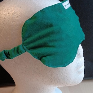 May include: A dark green sleep mask with an elasticated strap, displayed on a white mannequin head. The mask is leaf-shaped and features a small white tag with the text "Seven Green Cats".
