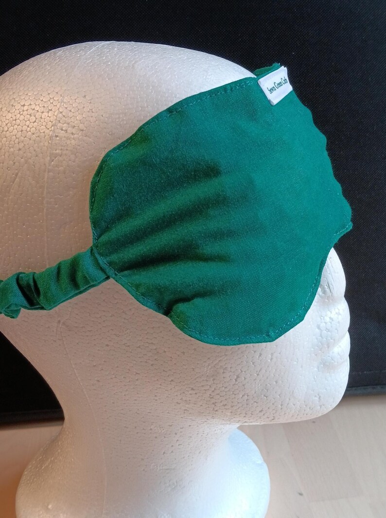 May include: A dark green sleep mask with an elasticated strap, displayed on a white mannequin head. The mask is leaf-shaped and features a small white tag with the text "Seven Green Cats".