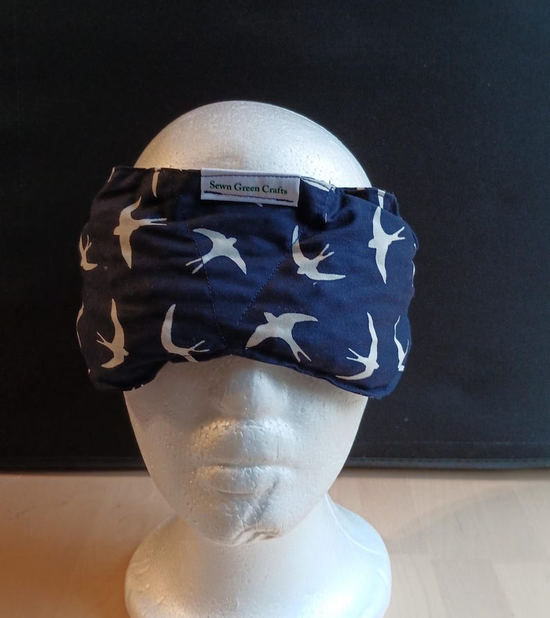 May include: A navy blue sleep mask featuring a white bird design. The mask is made of fabric and has a label that reads "Sewn Green Crafts". It is designed to block out light for improved sleep.