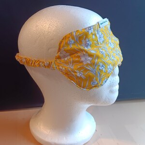 May include: A yellow sleep mask with a floral pattern of white and light blue flowers and leaves. The mask is on a white mannequin head. The elastic band is yellow. The label on the mask says "Sewn Green Life".