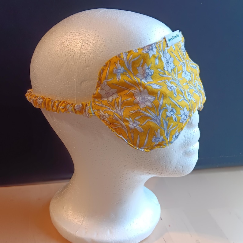May include: A yellow sleep mask with a floral pattern of white and light blue flowers and leaves. The mask is on a white mannequin head. The elastic band is yellow. The label on the mask says "Sewn Green Life".