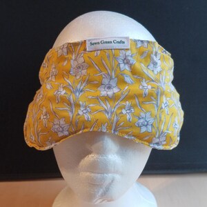 May include: A yellow floral headband with white and grey flowers. The headband is made of fabric and has the text "Sewn Green Crafts" on a small white tag. The headband is displayed on a white mannequin head.