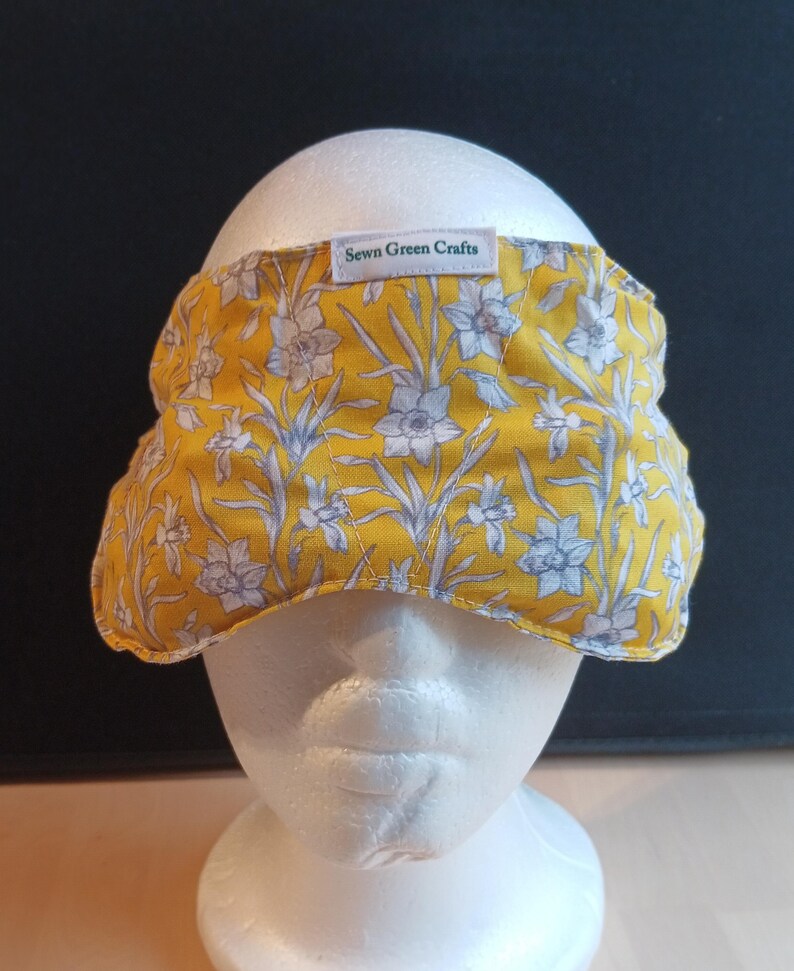 May include: A yellow floral headband with white and grey flowers. The headband is made of fabric and has the text "Sewn Green Crafts" on a small white tag. The headband is displayed on a white mannequin head.