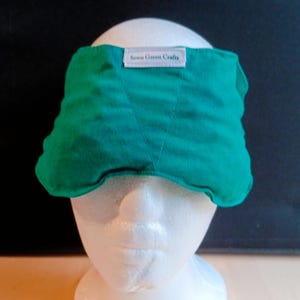 May include: A teal eye mask with a stitched design, worn on a white mannequin head. The mask has a label that reads "Sewn Green Crafts". The background is black.