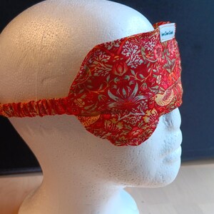 May include: A red sleep mask featuring a detailed floral and bird design in gold and green. The mask has an elasticated strap and a small white label that reads "Even Green Castle". The sleep mask is designed to block out light.