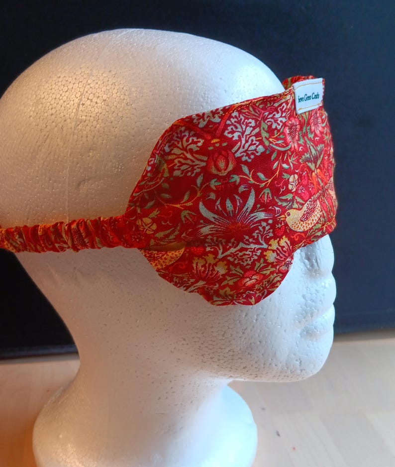 May include: A red sleep mask featuring a detailed floral and bird design in gold and green. The mask has an elasticated strap and a small white label that reads "Even Green Castle". The sleep mask is designed to block out light.