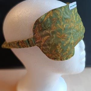 May include: A green and gold patterned sleep mask with a matching strap. The mask is made of fabric and has a label that reads "Sewn Green Crafts". The mask is designed to block out light.