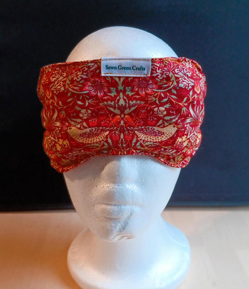 May include: A red and gold patterned headband with a floral and bird design. The fabric headband features a label that reads "Sewn Green Crafts". The headband is displayed on a white mannequin head.