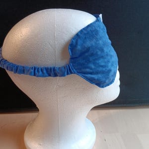 May include: A blue sleep mask with a textured pattern, designed to block out light. The mask is held in place by a blue elastic band. The sleep mask is displayed on a white mannequin head.