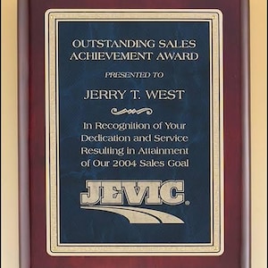 May include: A mahogany plaque with a gold trim and a blue plaque insert. The text on the plaque reads "Outstanding Sales Achievement Award" and "Presented to Jerry T. West". The text continues with "In Recognition of Your Dedication and Service Resulting in Attainment of Our 2004 Sales Goal". The plaque also features a silver logo with the text "JEVIC".