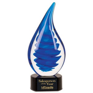 Op de afbeelding: Een blauwe en heldere glazen prijs in de vorm van een druppel, met een donkere basis. De prijs heeft een wervelend blauw ontwerp aan de binnenkant. De basis heeft de tekst "Salesperson of the Year" en "UPmedia CONFERENCE".