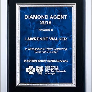 May include: A framed plaque with a blue marble-effect background. The text "DIAMOND AGENT 2018" is displayed in white, along with the name "LAWRENCE WALKER." The plaque is framed in black with a silver border, and includes the logos of Blue Cross Blue Shield.