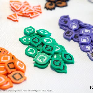 Puede incluir: Una colección de piezas de juego coloridas en varias formas y colores, incluyendo naranja, verde, morado y azul. Algunas piezas tienen diseños de hojas, mientras que otras tienen formas geométricas y números. Las piezas están dispersas sobre una superficie blanca.