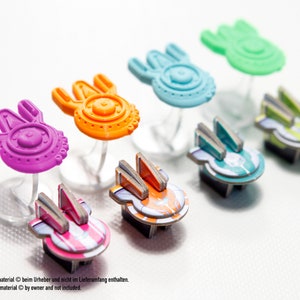 May include: Six colourful spaceship game pieces, each with a different design and colour. The pieces are made of plastic and are about 5 cm tall. The pieces are arranged in a row on a white surface.