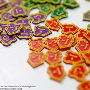 May include: A collection of colorful game tokens in various colors, including purple, red, green, and gray. Each token is shield-shaped with a gold border and features a number, such as -1 or 3. The tokens are scattered on a white surface.