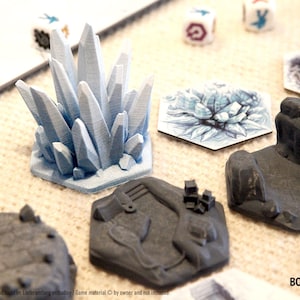Può includere: Una collezione di componenti di gioco, tra cui una grande formazione di cristalli azzurri, formazioni rocciose grigie e tessere esagonali con illustrazioni. Diversi dadi con simboli sono sparsi.
