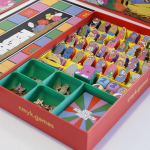 Può includere: Un set di gioco da tavolo con una scatola rossa dotata di scomparti interni contenenti numerosi piccoli gettoni personaggio colorati, dadi numerati e pezzi a forma di stella. Un tabellone di gioco con un percorso di caselle numerate etichettate "HIGER RASEN" è visibile accanto alla scatola, che riporta il testo "cmyk.games" sul lato.