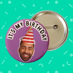 May include: Purple button with the text "It's My Birthday" in white. A man's face is in the centre of the button with a red and white striped party hat on his head.