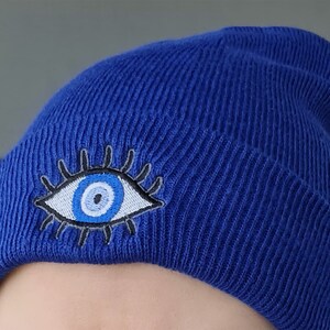 May include: A royal blue knitted beanie with an embroidered evil eye design in white, blue, and gray.