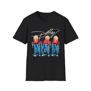 May include: A black short-sleeve t-shirt featuring a graphic print of Dolly Parton in three different poses. The design shows her wearing a red long-sleeve shirt and blue jeans, with the text Dolly Parton and Here You Come Again printed above.