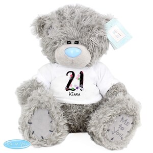 May include: Grey plush teddy bear wearing a white t-shirt with the number 21 and the name Kiara printed on it. The bear has a blue nose and is sitting on a white background. The bear is a Me to You bear.