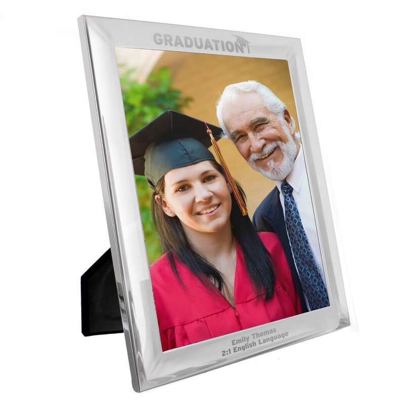 Pu&ograve; includere: Cornice per foto argentata con un design a forma di cappello di laurea. La cornice contiene una foto di una donna con un cappello e una toga da laurea con un uomo. Il testo sulla cornice recita "GRADUATION" e "Emily Thomas 2:1 English Language".