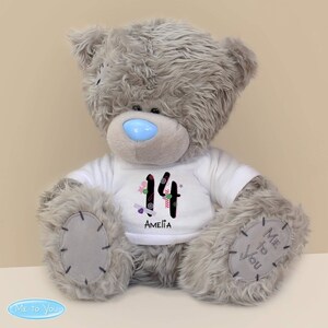 May include: Grey plush teddy bear wearing a white t-shirt with the number 14 decorated with colourful candy and the name Amelia. The bear has a blue nose and the words "Me to You" are printed on the bottom of the image.