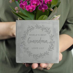 May include: Grey concrete planter with a floral wreath design and the text "World's Best Grandma Love Lily".