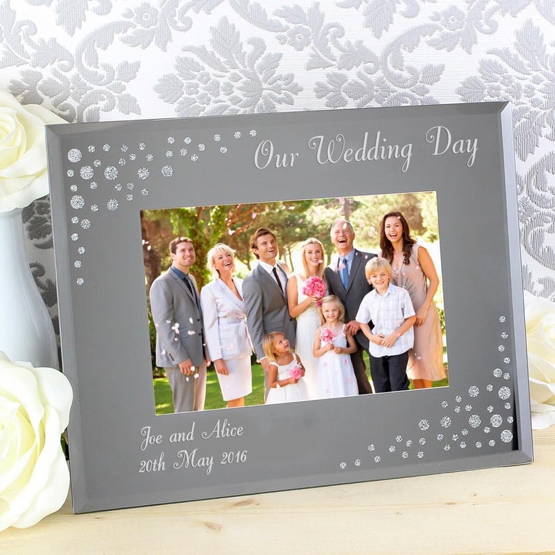 K&ouml;nnte beinhalten: Ein silberner Bilderrahmen mit wei&szlig;em Hintergrund und einem Foto einer Hochzeitsgesellschaft. Der Rahmen hat einen silbernen Glitzer-Rand und der Text "Our Wedding Day" und "Joe and Alice 20th May 2016" ist in den Rahmen graviert.
