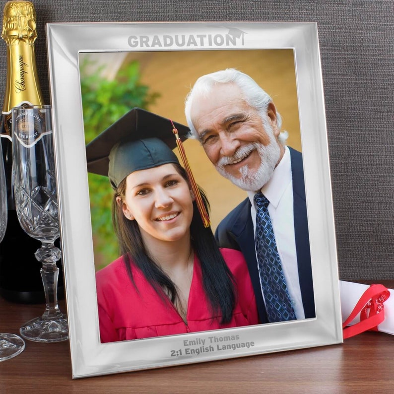 Pu&ograve; includere: Cornice per foto d'argento per la laurea con una foto di un laureato e un membro della sua famiglia. La cornice ha le parole "GRADUATION" incise in alto e il nome del laureato, il titolo di studio e l'anno di laurea incisi in basso.