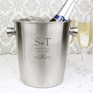 May include: Silver champagne bucket with a personalized inscription: "S.T. Greenway Hall 28th May 2018". The bucket is filled with ice and a bottle of champagne is inside.