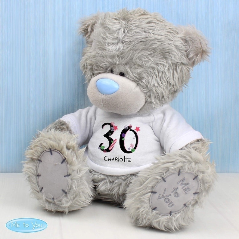 May include: A grey plush teddy bear wearing a white t-shirt with the number 30 and the name Charlotte printed on it. The bear is sitting on a light blue background.