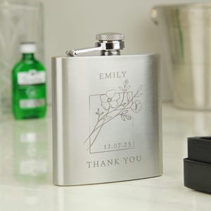 May include: Silver stainless steel hip flask with an engraved floral design, the name "EMILY", the date "12.07.23", and the words "THANK YOU".