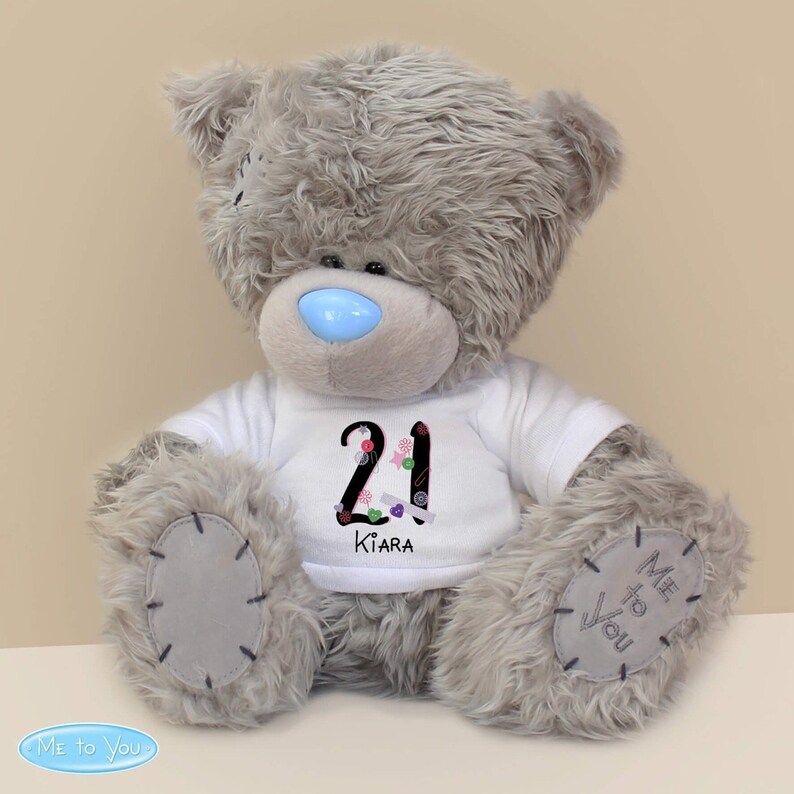 May include: A grey plush teddy bear wearing a white t-shirt with the number 21 and the name Kiara printed on it. The bear has a blue nose and the words "Me to You" are printed on its paw.
