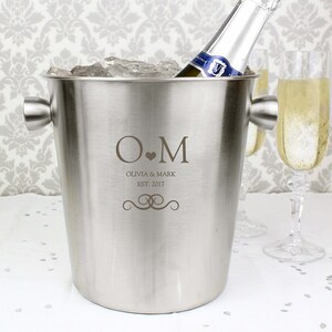 May include: A silver ice bucket with a personalized inscription that reads "O.M Olivia & Mark Est. 2017". The bucket is filled with ice and a bottle of champagne is placed inside.