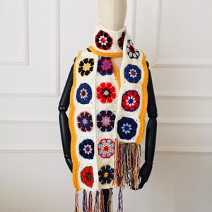 May include: A handmade crochet scarf featuring a floral pattern in red, blue, yellow, and purple. The scarf has a cream-colored background with a yellow border and colorful fringe.
