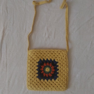May include: A yellow crocheted crossbody bag with a colorful granny square design. The bag has a long strap and is made with a textured yarn.