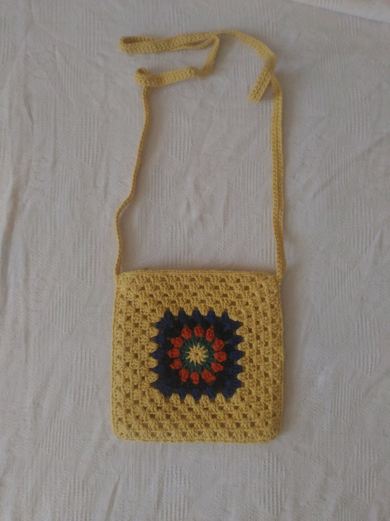 May include: A yellow crocheted crossbody bag with a colorful granny square design. The bag has a long strap and is made with a textured yarn.