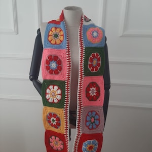 Puede incluir: Una larga bufanda de ganchillo con un estampado floral colorido. La bufanda está hecha de cuadrados con flores de diferentes colores en el centro. La bufanda tiene flecos blancos en los extremos.