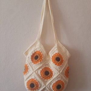May include: Cream-colored crochet tote bag featuring a sunflower design. The bag is made of squares with orange and brown sunflower motifs. The bag has a long strap for carrying over the shoulder.