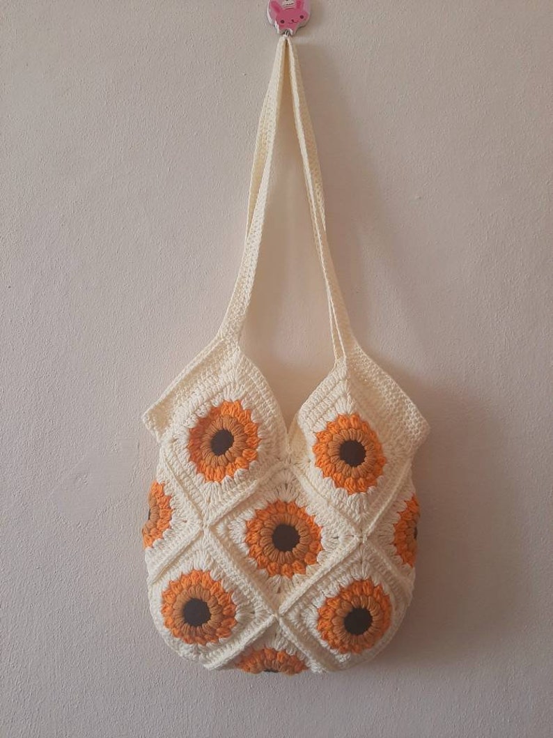 May include: Cream-colored crochet tote bag featuring a sunflower design. The bag is made of squares with orange and brown sunflower motifs. The bag has a long strap for carrying over the shoulder.