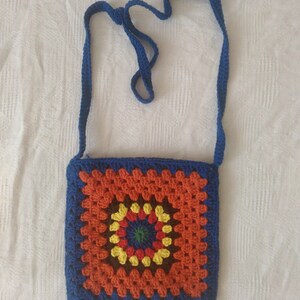 May include: A crocheted crossbody bag with a blue strap and a colorful granny square design. The square features a circular pattern in shades of orange, yellow, green, brown, and red.